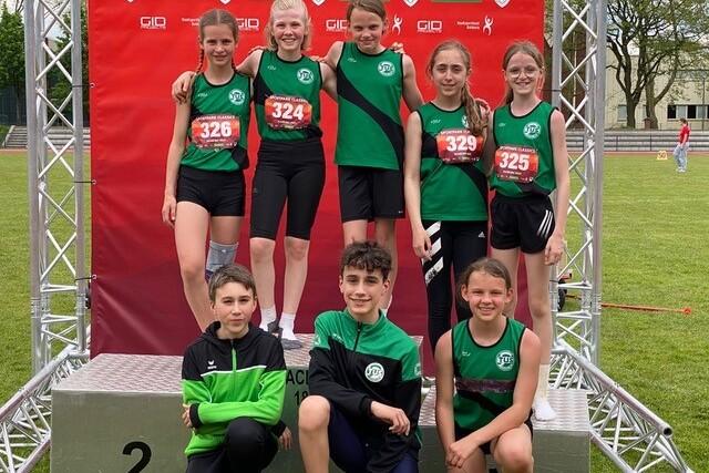 Gruppe von neun jungen Athleten in Sportkleidung vor einem roten Hintergrund mit Startnummern und Medaillen.