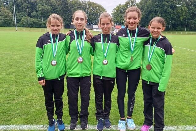 F&uuml;nf M&auml;dchen in gr&uuml;nen Sportjacken mit Medaillen stehen auf einem Fu&szlig;ballfeld und l&auml;cheln in die Kamera.