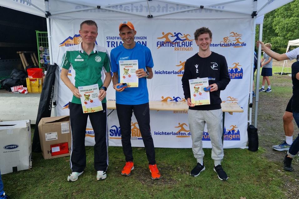 Drei M&auml;nner stehen unter einem Zelt, halten Urkunden in den H&auml;nden und tragen Sportswear nach einem Wettkampf.