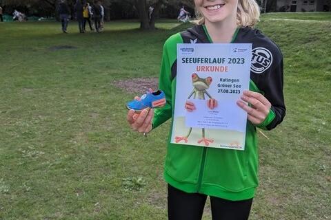 M&auml;dchen h&auml;lt Urkunde und Medaille vom Seefuferlauf 2023 in einer Parklandschaft. Sie tr&auml;gt einen gr&uuml;nen Sportjacke.