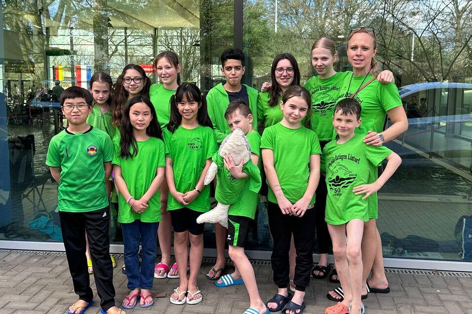 Gruppe von 14 Kindern und Jugendlichen in gr&uuml;nen T-Shirts, stehend vor gro&szlig;en Fenstern eines Schwimmbades.