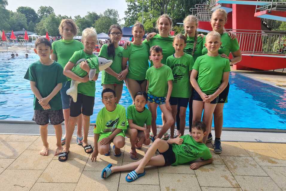 Gruppenzusammenkunft von Kindern in gr&uuml;nen T-Shirts am Schwimmbeckenrand, vor Sprungturm und Wasser.