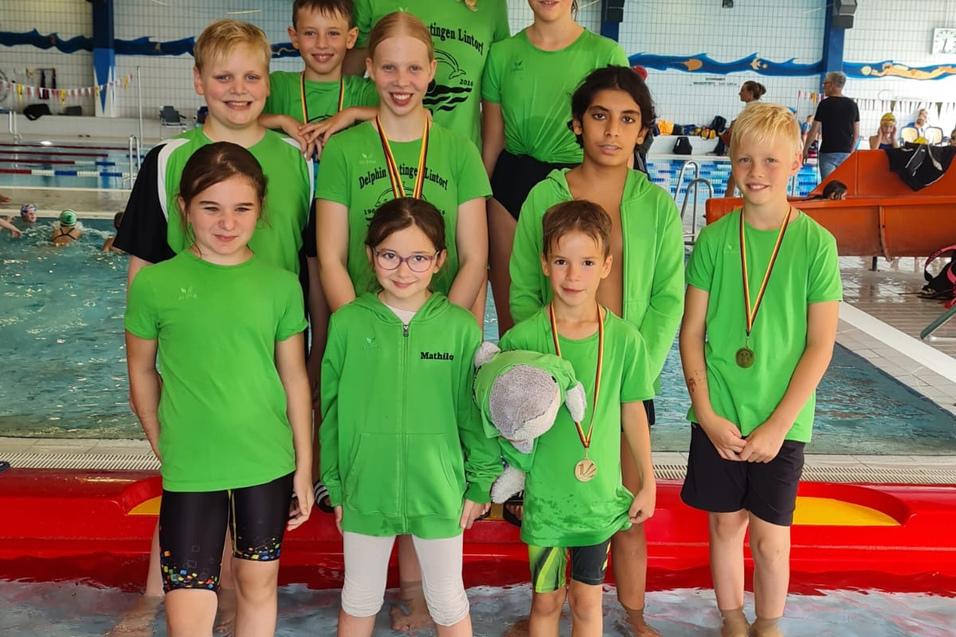 Gruppe von Schwimmern in gr&uuml;nen T-Shirts mit Medaillen, posierend im Schwimmbad. Hintergr&uuml;nde zeigen Wasser und andere Schwimmer.