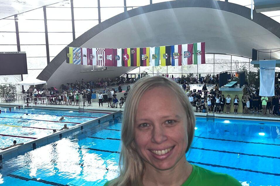 Frau mit blonden Haaren in gr&uuml;nem Shirt l&auml;chelt vor einem Schwimmbecken mit Zuschauern und Fahnen im Hintergrund.