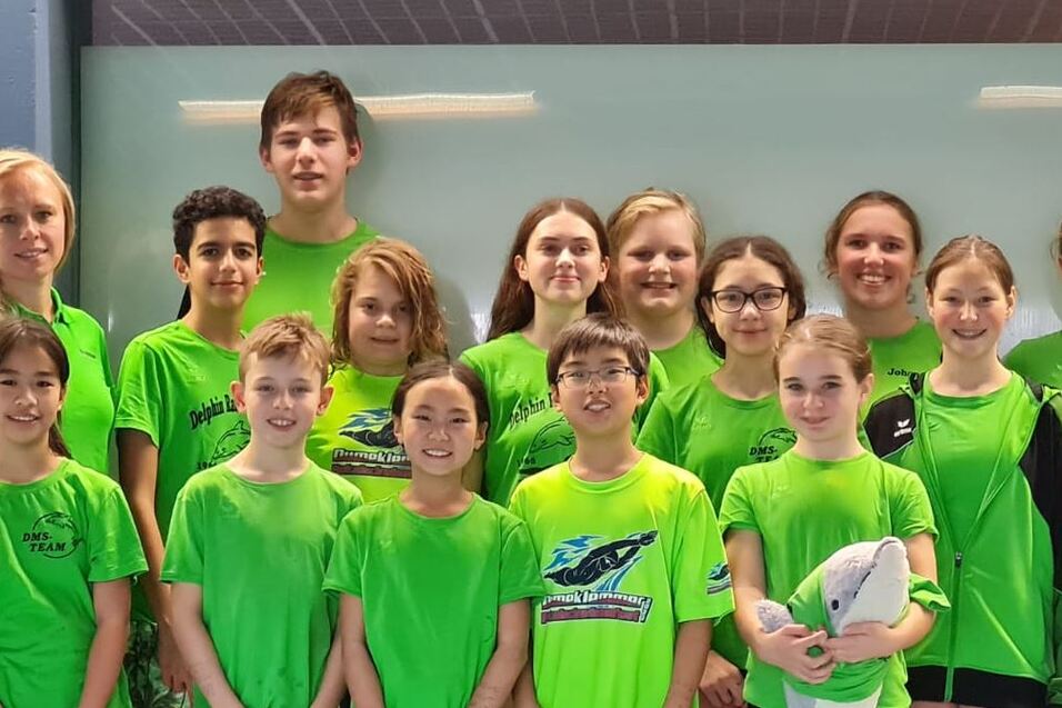 Gruppe von 16 Jugendlichen in gr&uuml;nen T-Shirts, versammelt in einer Sporthalle vor einem hellen Hintergrund.