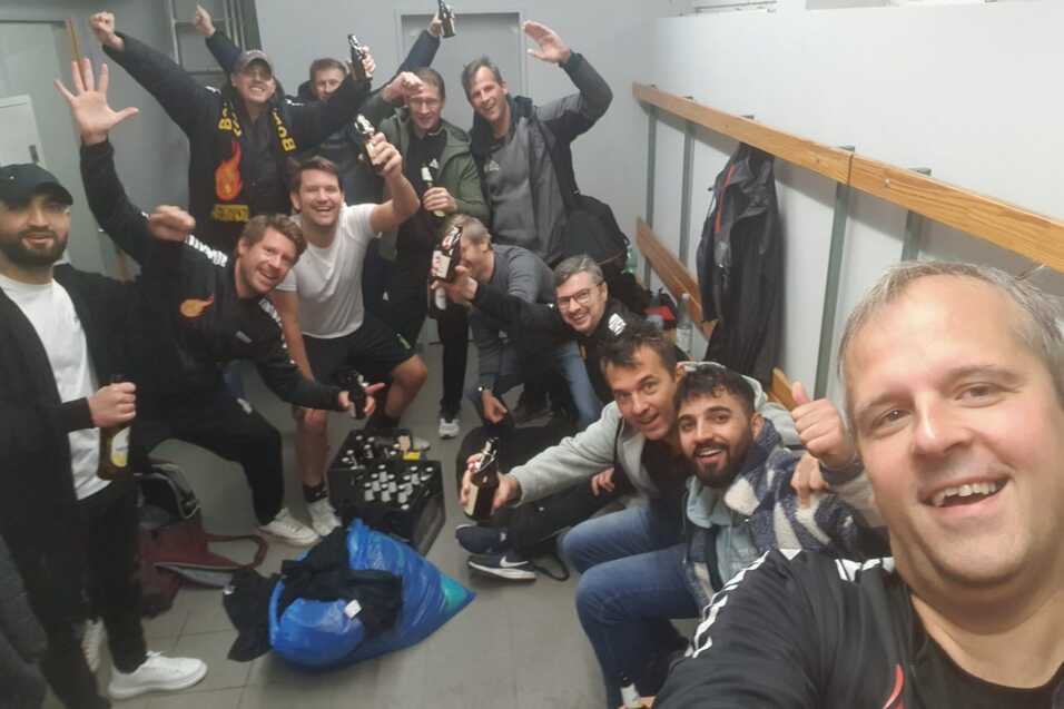 Gruppenfoto von zehn M&auml;nnern in Sportkleidung in einem Umkleideraum, l&auml;chelnd und mit Getr&auml;nken in der Hand.