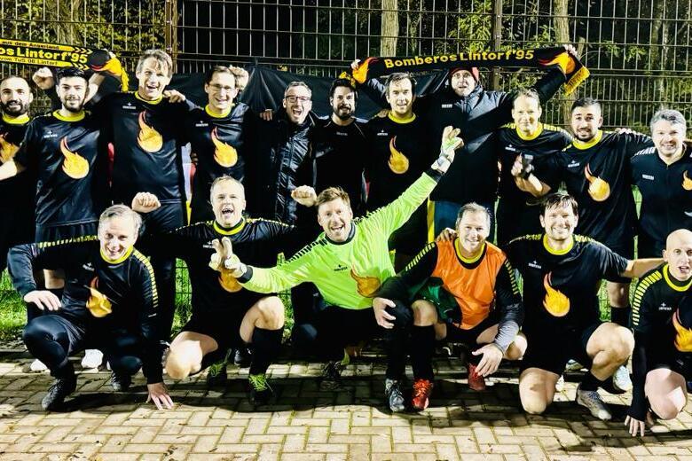 Gruppenfoto einer Fu&szlig;ballmannschaft in schwarzen Trikots, mit Schals und freudigem Posieren auf einem Sportplatz.