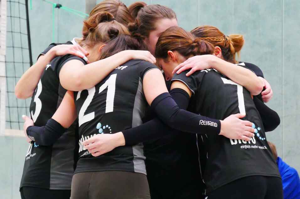 Sechs Volleyballspielerinnen umarmen sich in einer Sporthalle, alle in schwarzen Trikots und gr&uuml;nen Socken.
