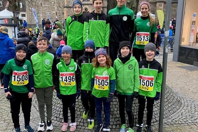 Gruppe von Kindern in gr&uuml;nen Sportjacken mit Startnummern beim Lauf, stehen drau&szlig;en auf einem Platz.