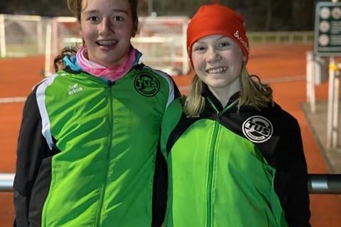 Zwei l&auml;chelnde M&auml;dchen in gr&uuml;nen Sportjacken stehen bei Nacht an einem Sportplatz mit Fu&szlig;balltoren im Hintergrund.