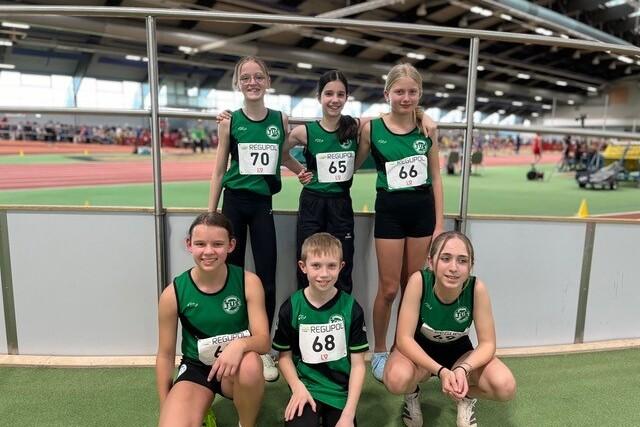 Sechs junge Leichtathleten in gr&uuml;nen Trikots posieren l&auml;chelnd vor einer Sporthalle.