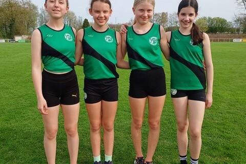 Vier junge Sportlerinnen in einheitlicher gr&uuml;ner Sportbekleidung stehen l&auml;chelnd auf einem gr&uuml;nen Rasenfeld.