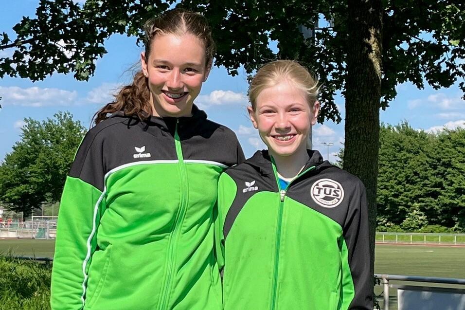 Zwei junge Sportlerinnen in gr&uuml;nen Jacken stehen l&auml;chelnd vor einem Baum auf einer Wiese.