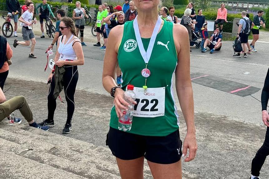 Frau in Sportkleidung mit Medaille und Wasserflasche steht nach einem Lauf im Park, umgeben von anderen Teilnehmern.