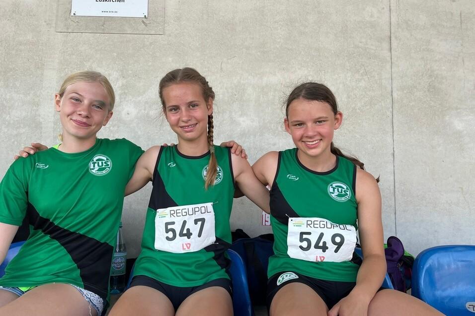 Drei Sportlerinnen in gr&uuml;nen Trikots sitzen nebeneinander auf einer Trib&uuml;ne und l&auml;cheln in die Kamera.