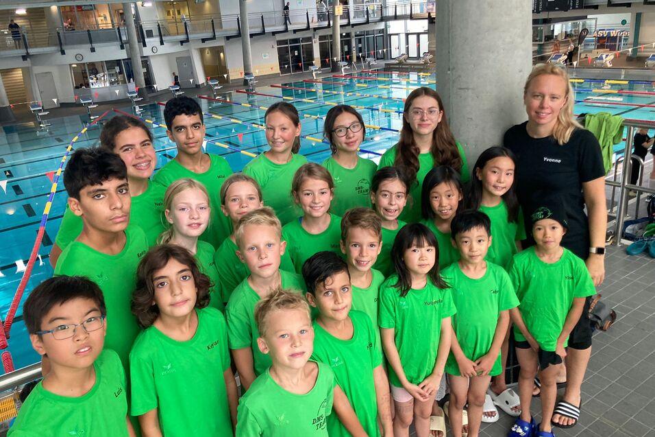 Gruppe von Schwimmsportlern in gr&uuml;nen T-Shirts posiert vor einem Schwimmbecken mit mehreren Bahnen.
