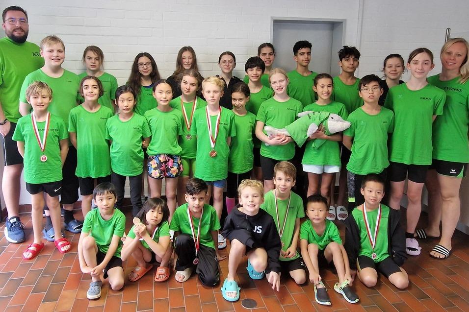Gruppe von Kindern und Jugendlichen in gr&uuml;nen T-Shirts mit Medaillen, umgeben von Betreuern in Sportumgebung.