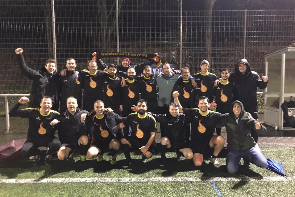 Mannschaft von Fu&szlig;ballspielern posiert in schwarzen Trikots mit Flammenlogo auf einem Kunstrasenplatz bei Nacht.
