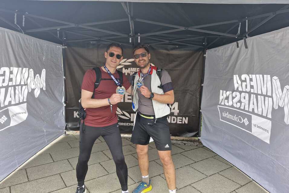 Zwei Sportler posieren unter einem Zeltdach und halten Medaillen in den H&auml;nden. Sie tragen Sportkleidung und Rucks&auml;cke.