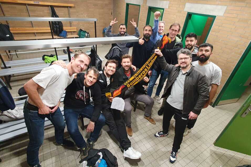 Gruppe von zehn M&auml;nnern in einem Umkleideraum, l&auml;chelnd und posierend mit einem Schal in den H&auml;nden.