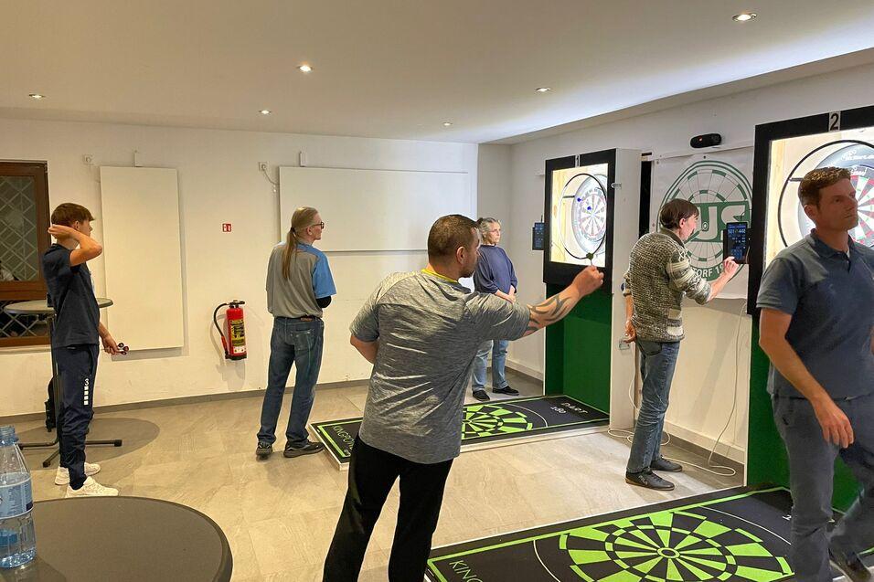 Gruppe von fünf Personen beim Darten in einem hellen Raum mit zwei Dartscheiben und Grünflächen.
