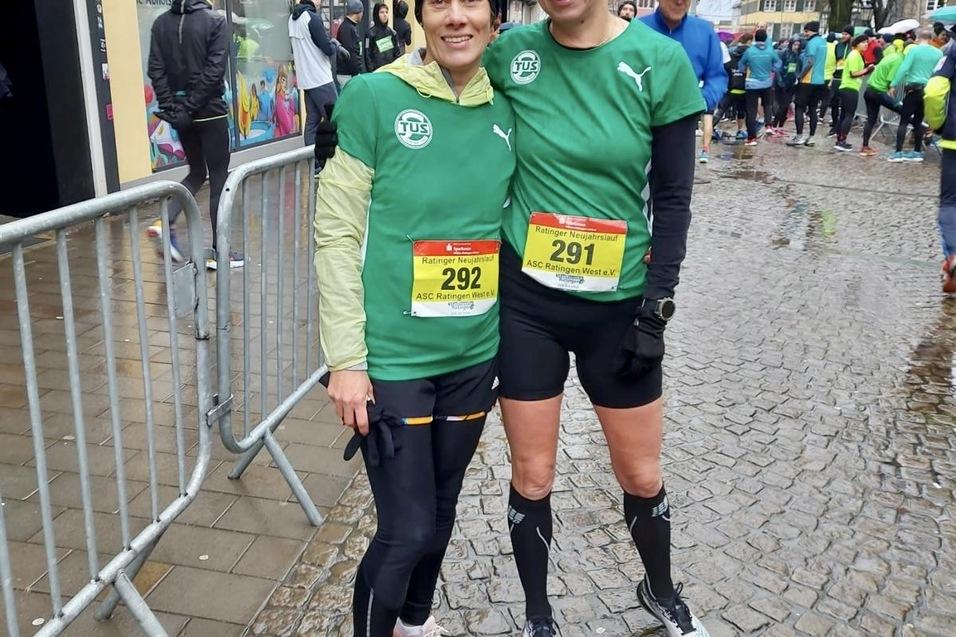 Zwei Frauen in gr&uuml;nen Laufshirts mit Startnummern stehen auf einer nassen Stra&szlig;e vor einer Laufveranstaltung.