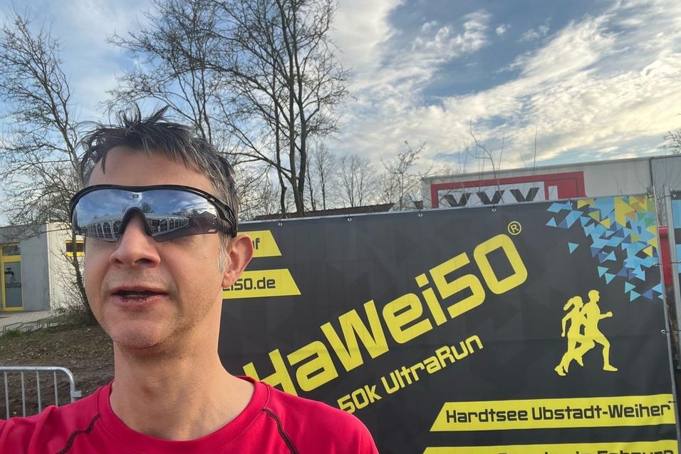 Mann in rotem Shirt und Sonnenbrille posiert vor einem Banner des HaWei50 UltraRuns. Hintergrund mit B&auml;umen.