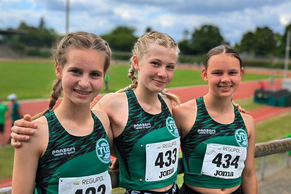 Drei junge Athletinnen in Sportkleidung posieren mit Startnummern auf einer Leichtathletikanlage bei strahlendem Himmel.