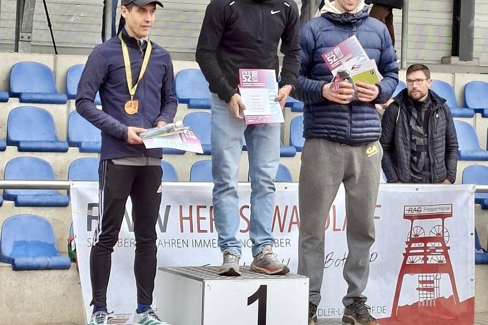 Drei Läufer stehen auf dem Siegerpodest, halten Urkunden und Medaillen, mit Zuschauern im Hintergrund.