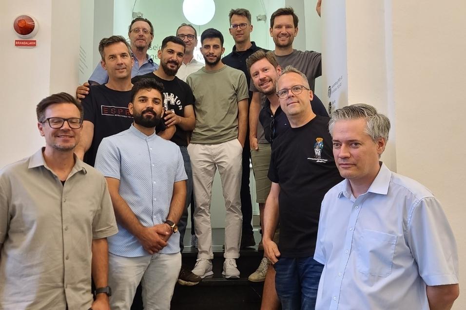 Gruppenfoto von zw&ouml;lf M&auml;nnern, die auf einer Treppe stehen, mit modernen Innenr&auml;umen und Deckenleuchten im Hintergrund.