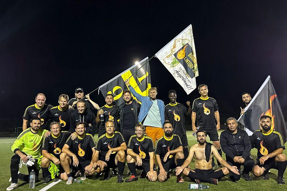 Mannschaft von 20 Fu&szlig;ballspielern in schwarzer Kleidung posiert mit Fahnen auf einem Kunstrasenplatz bei Nacht.