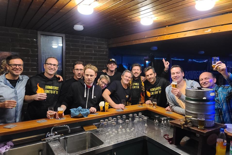 Gruppe von zw&ouml;lf Personen l&auml;chelt an einer Bar, h&auml;lt Gl&auml;ser und posiert vor einem Bierfass.