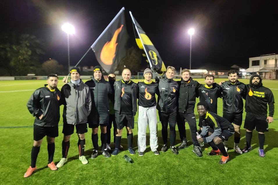 Gruppenshot von 11 Fu&szlig;ballspielern in einheitlicher Sportkleidung, die Flaggen auf einem beleuchteten Feld schwenken.