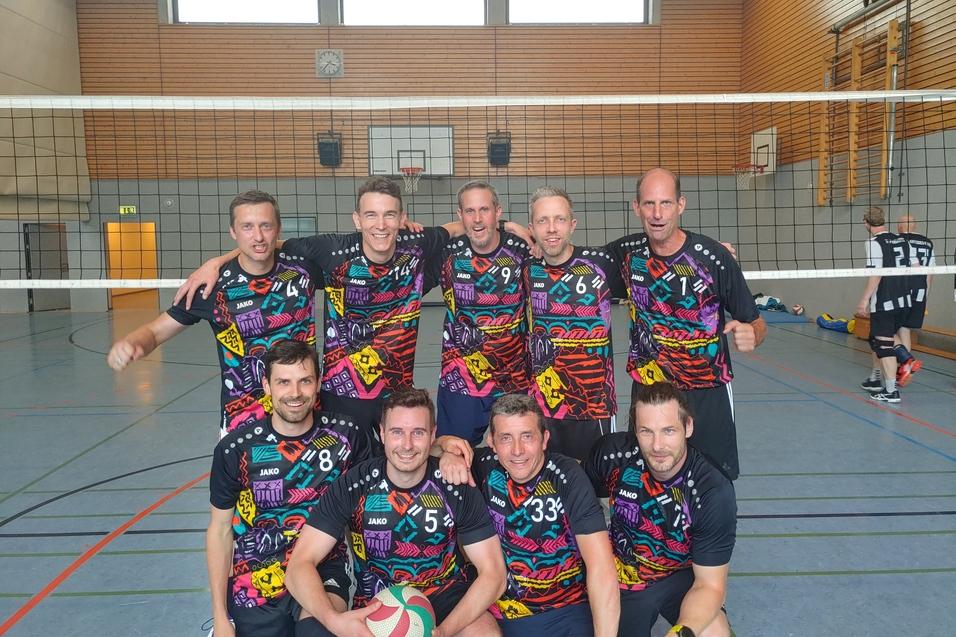 Gruppe von acht männlichen Volleyballspielern in bunten Trikots, posierend vor einem Volleyballnetz in einer Sporthalle.