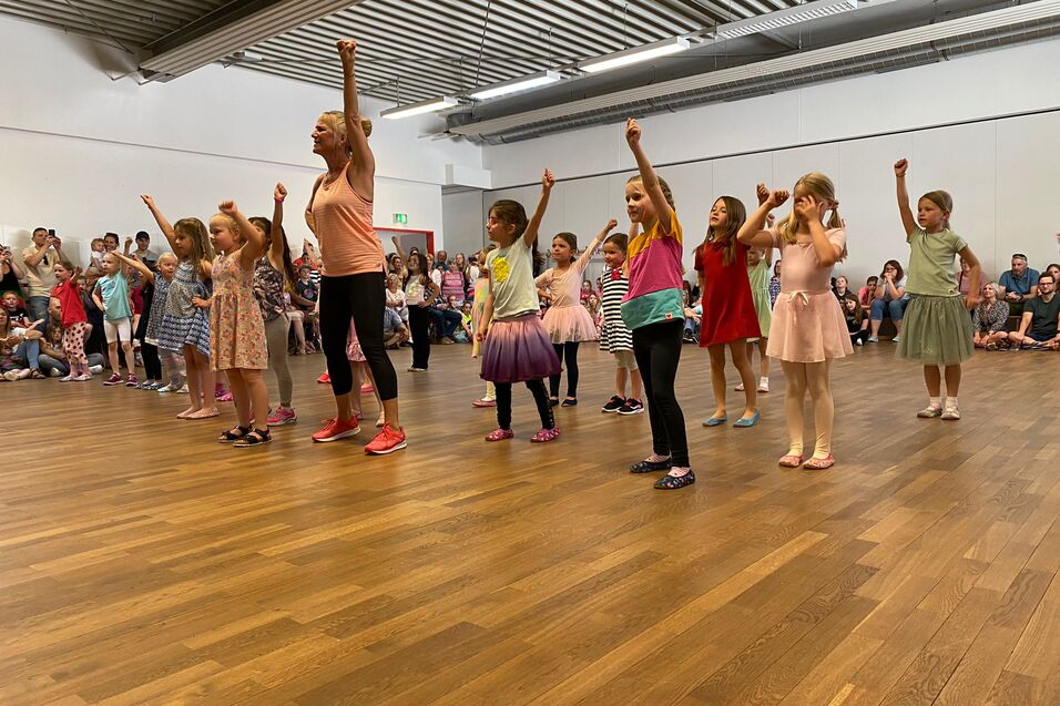 Gruppe von M&auml;dchen in bunten Outfits tanzt auf einer B&uuml;hne, w&auml;hrend Eltern im Hintergrund zuschauen.