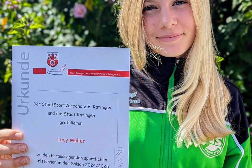 Mädchen in grüner Sportjacke hält Urkunde mit dem Text „Lucy Müller“ vor einer blühenden Hintergrundpflanze.