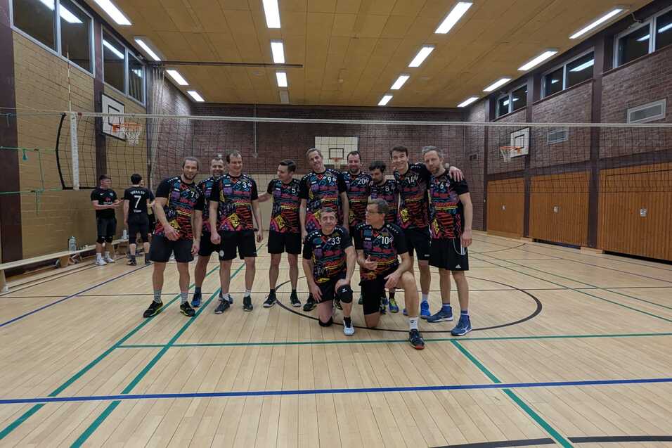 Gruppe von zehn Volleyballspielern in Sportkleidung posiert l&auml;chelnd in einer Halle mit Volleyballnetz im Hintergrund.