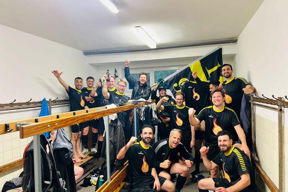 Gruppenshot von Spielern in schwarzen Trikots, die in einer Umkleidekabine jubeln und eine Flagge halten.