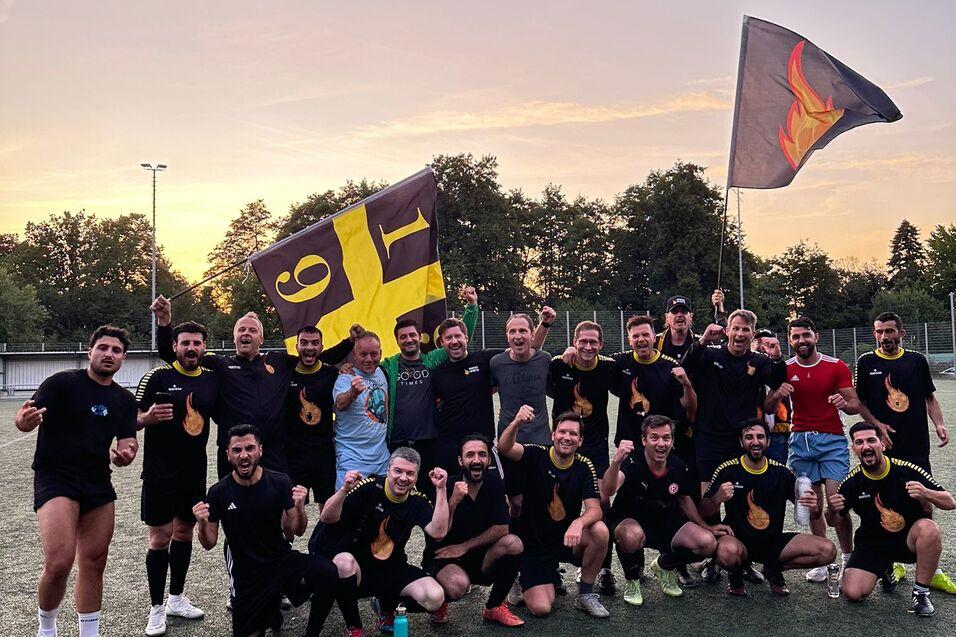 Gruppe von Fu&szlig;ballspielern in schwarzen Trikots mit einer Fahne auf einem Sportplatz bei Sonnenuntergang.
