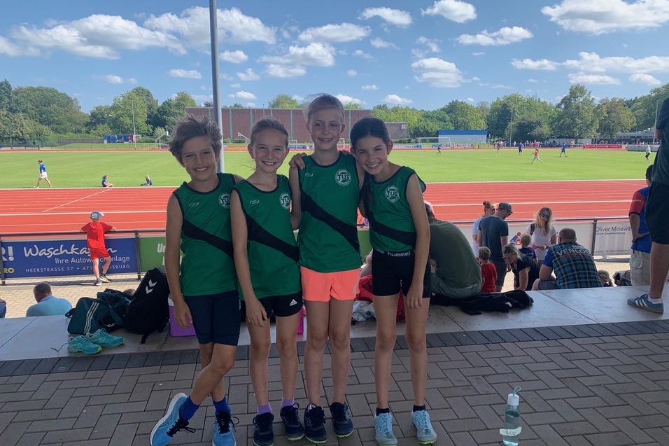 Vier Kinder in gr&uuml;nen Sporttrikots stehen l&auml;chelnd auf einer Trib&uuml;ne mit einer Laufbahn im Hintergrund.