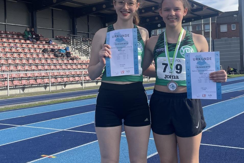 Zwei junge Athletinnen in Sportkleidung halten Urkunden und Medaille auf einer blauen Leichtathletikbahn w&auml;hrend eines Wettkampfs.
