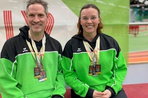 Zwei Sportler in gr&uuml;nen Trikots mit Medaillen sitzen l&auml;chelnd auf einer Trib&uuml;ne in einer Sporthalle.