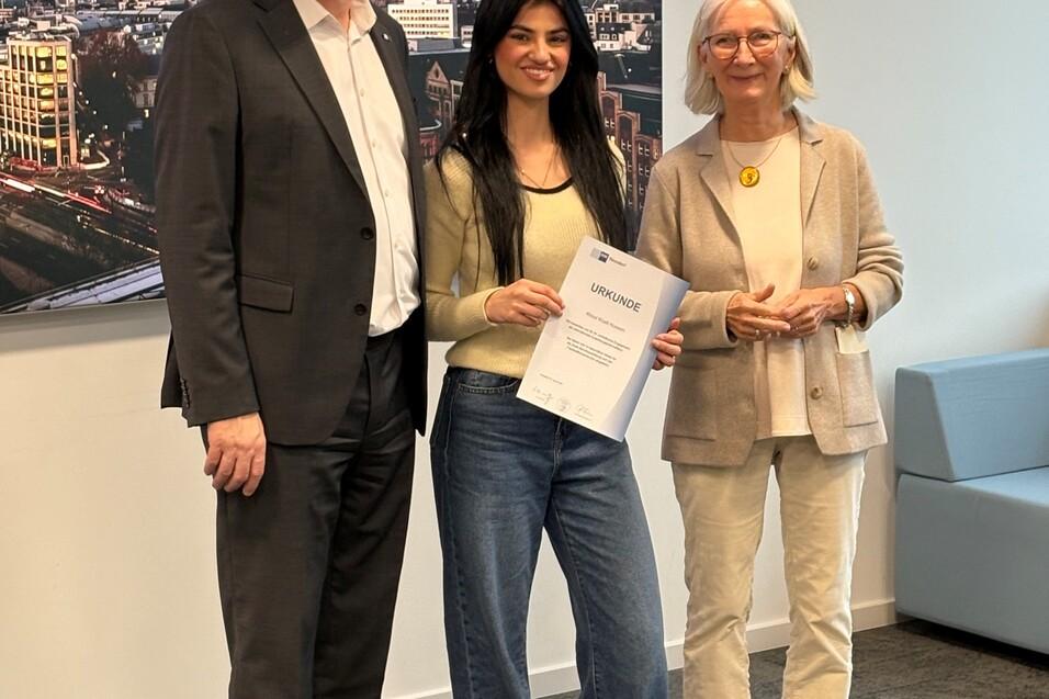 Drei Personen stehen in einem B&uuml;ro, halten ein Zertifikat und l&auml;cheln vor einer Stadtansicht im Hintergrund.