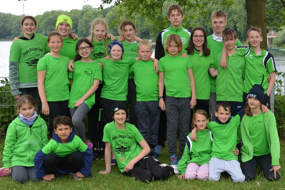 Gruppe von Jugendlichen in gr&uuml;nen T-Shirts steht am Ufer eines Gew&auml;ssers, umgeben von B&auml;umen.