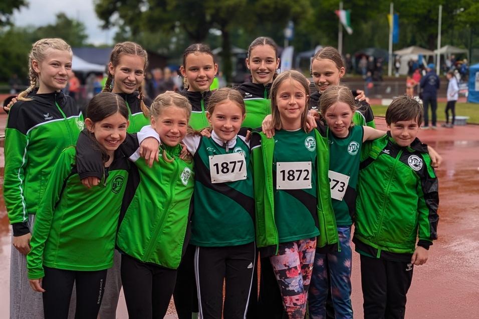 Gruppe von zehn jungen Sportlern in grünen Trikots und Jacken, posierend auf einem nassen Sportgelände.
