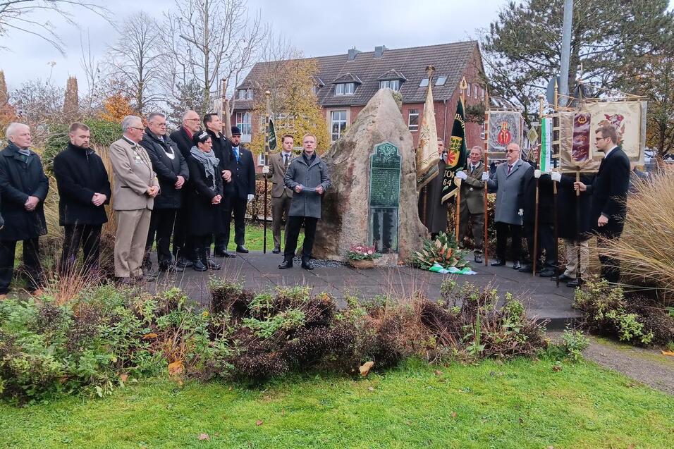 Menschen stehen versammelt vor einem Denkmal, umgeben von Pflanzen und Herbstwald, bei einer Gedenkveranstaltung.