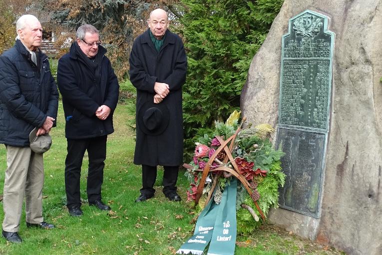 Drei Personen stehen vor einem Gedenkstein, w&auml;hrend sie eine Kranzniederlegung im Freien durchf&uuml;hren.