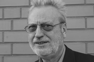 Portr&auml;t eines &auml;lteren Mannes mit Brille und grauem Bart vor einer Backsteinmauer.