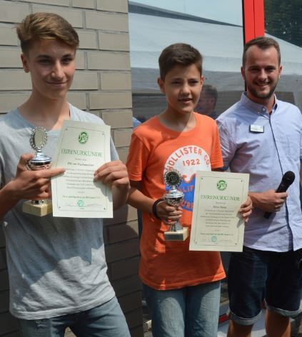 Zwei junge Gewinner halten Pokale und Urkunden, w&auml;hrend ein Moderator mit Mikrofon neben ihnen steht.