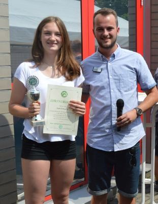 Zwei Personen stehen vor einem Geb&auml;ude: Ein M&auml;dchen h&auml;lt eine Troph&auml;e und ein Zertifikat, ein Junge spricht ins Mikrofon.